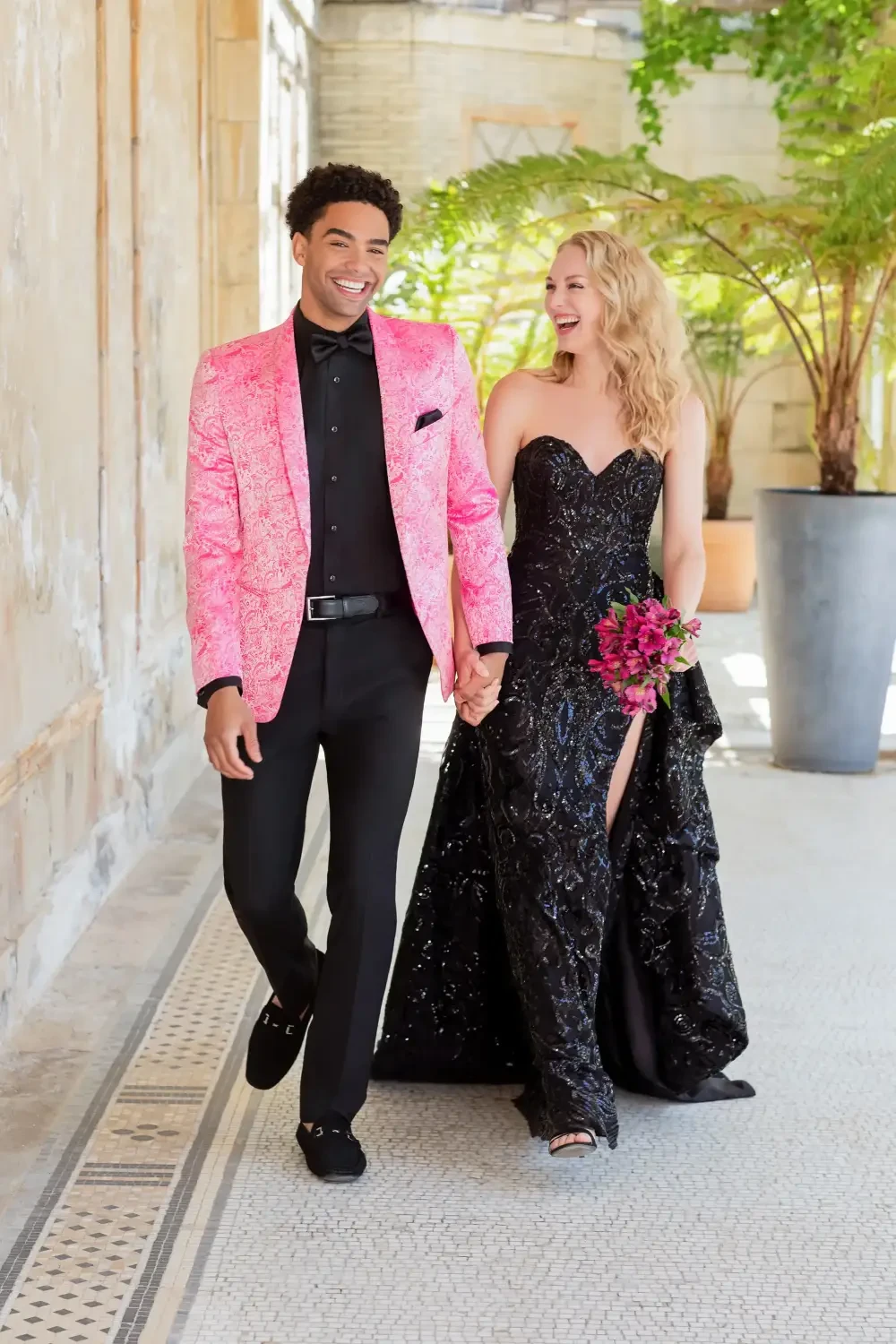 A couple walks hand in hand, the man wearing a bright pink jacket and black pants, and the woman in a black strapless gown with a front slit, holding a bouquet of flowers.