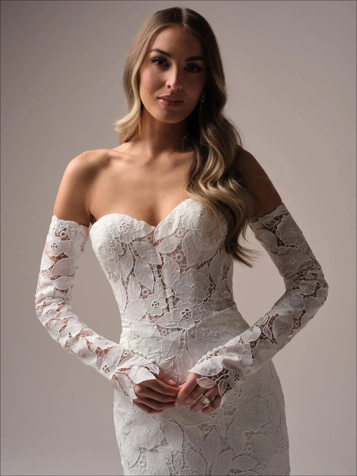 Model is wearing a strapless boho mermaid wedding gown with off-the-shoulder lace gauntlet sleeves and allover lace.