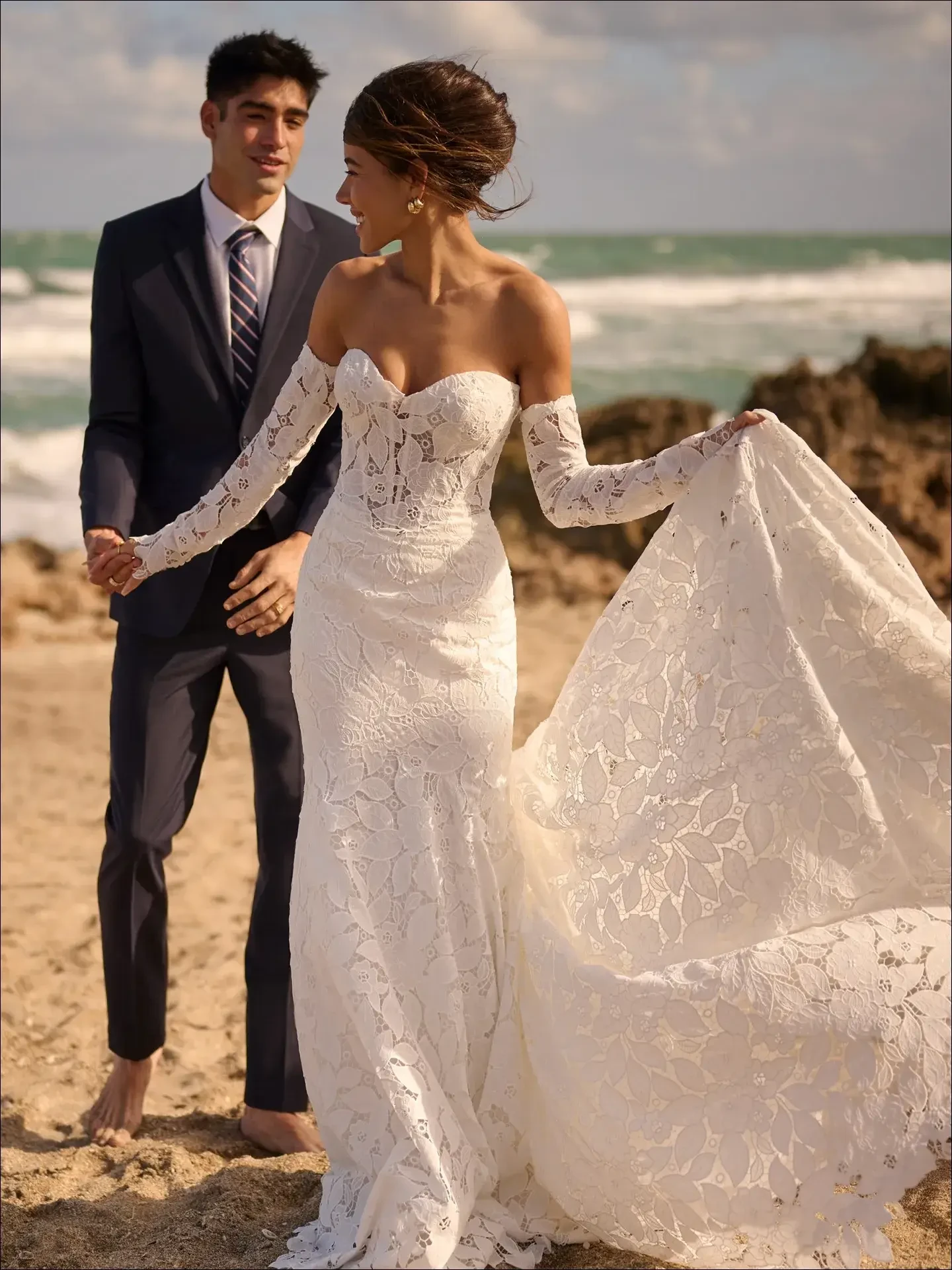 A joyful bride and groom walk barefoot on a sandy beach. She wears a lace wedding dress with flowing sleeves, while he dons a navy suit, ocean waves behind.