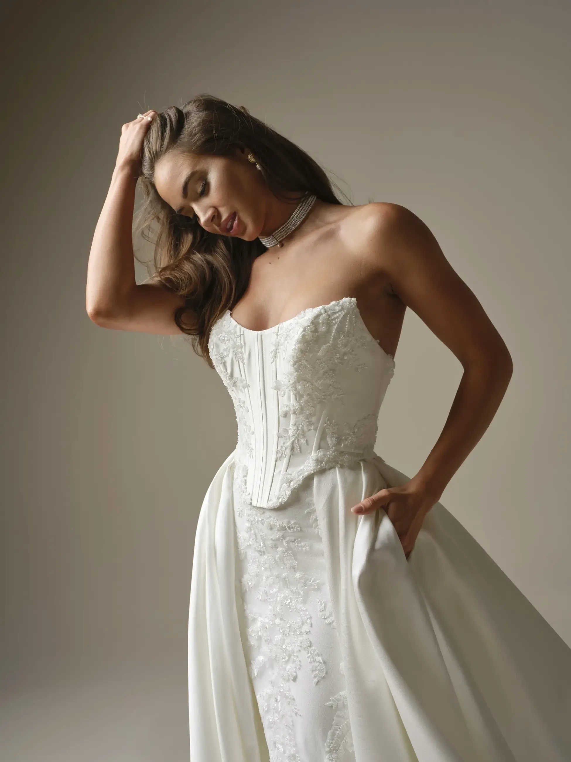 Bride in an elegant strapless white gown with intricate lace and a flowing skirt. She gazes down, hand in hair, exuding grace and sophistication.