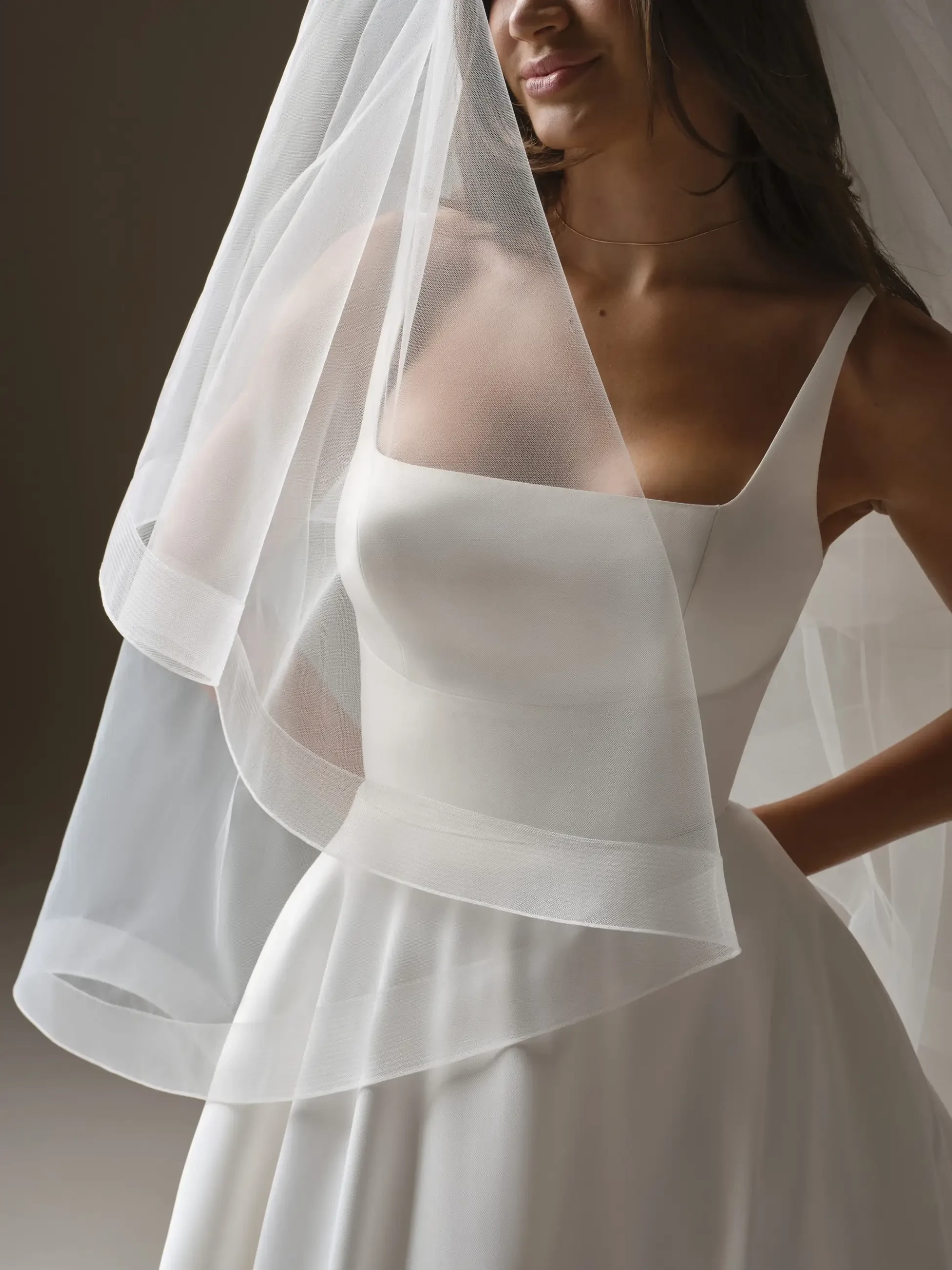 A bride in a white gown with spaghetti straps smiles softly. A delicate, flowing veil partially covers her face, creating a romantic and serene atmosphere.