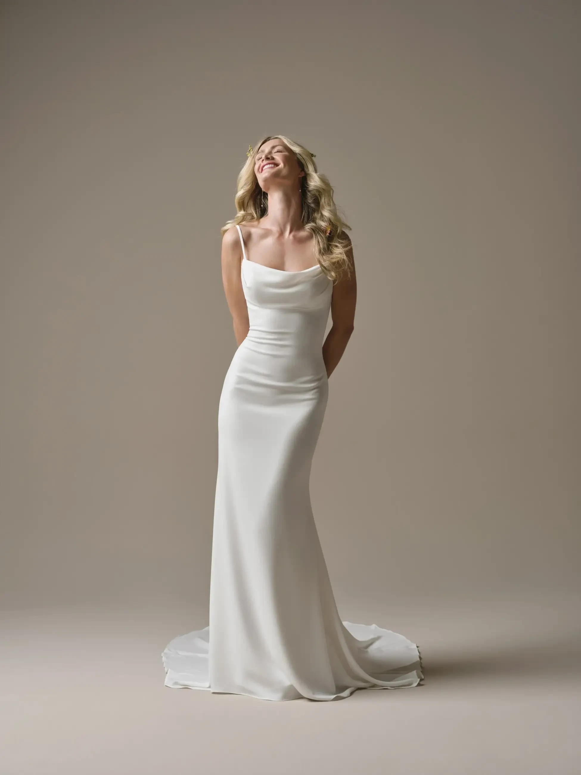 A woman in a white wedding dress stands with her back slightly arched and her hands clasped behind her. She is smiling and looking upwards against a neutral background.