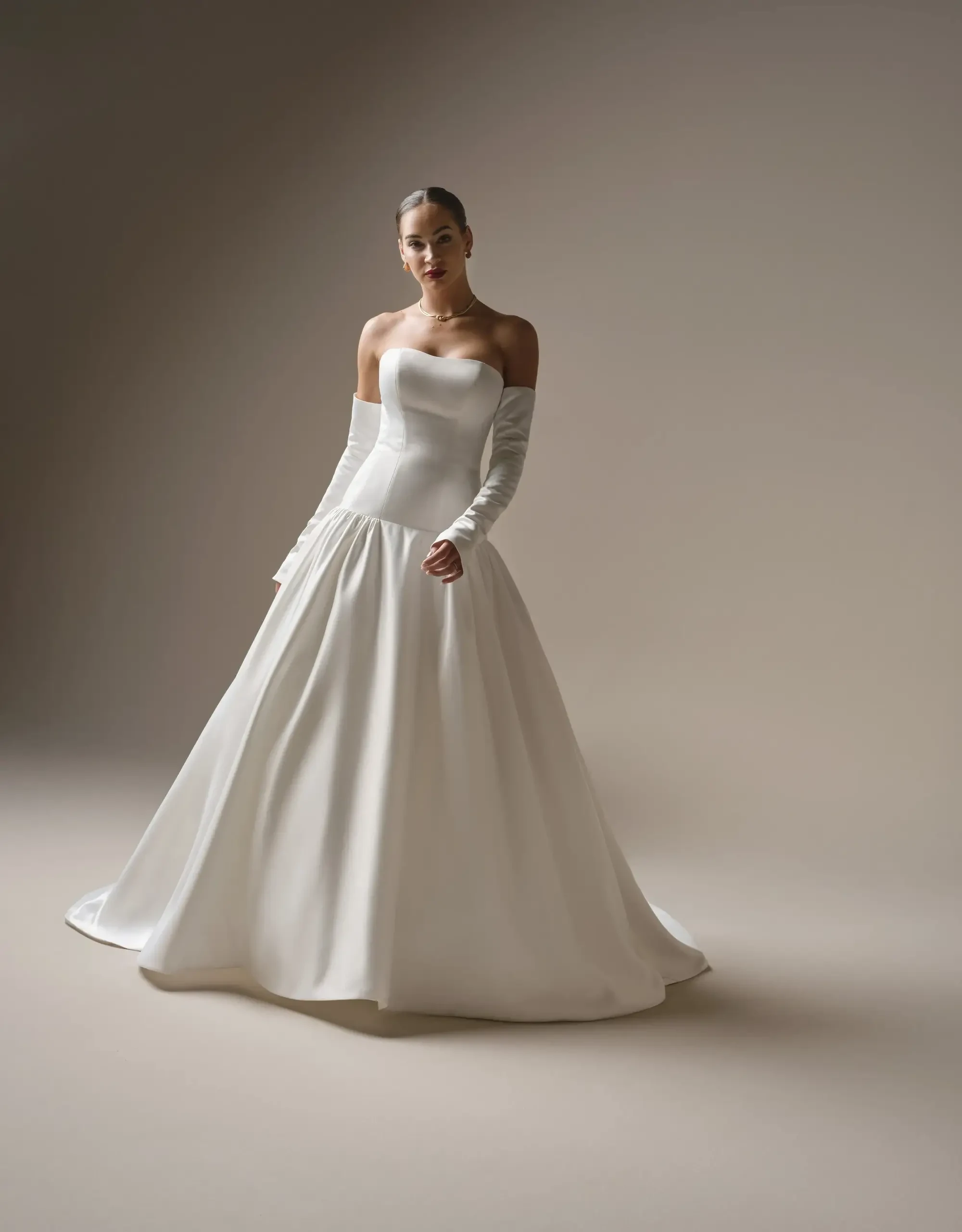 A woman in a strapless, white wedding gown with long sleeves, posing against a neutral background.