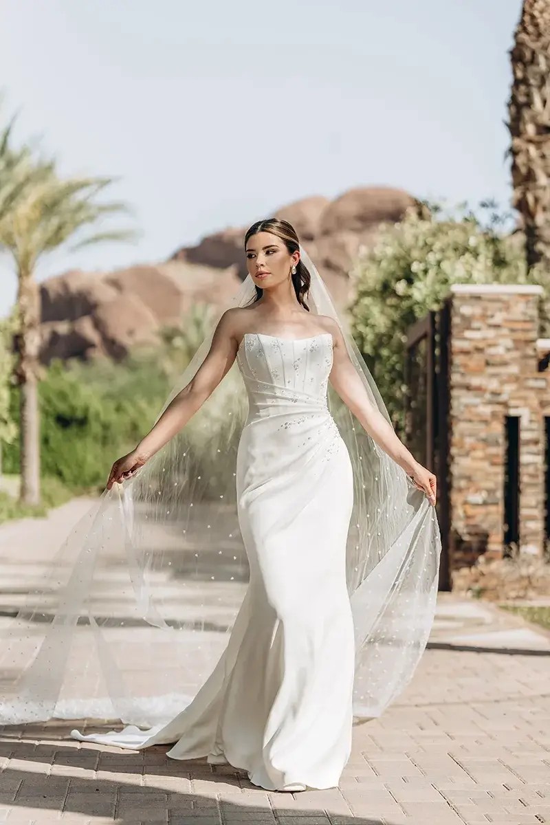 Model is wearing a column wedding dress with a strapless bodice and 3D floral accents