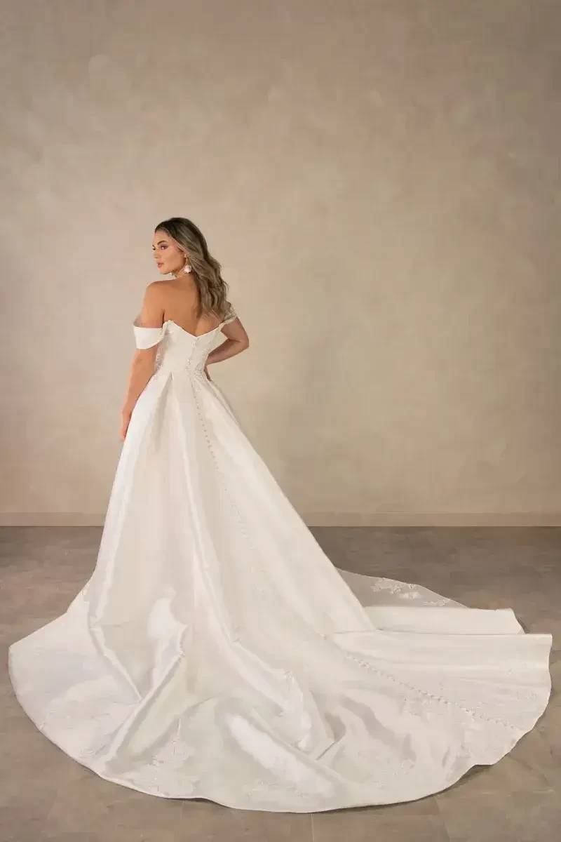 A woman wearing a white off-the-shoulder gown stands with her back to the camera, showcasing the dress's long train against a neutral background.