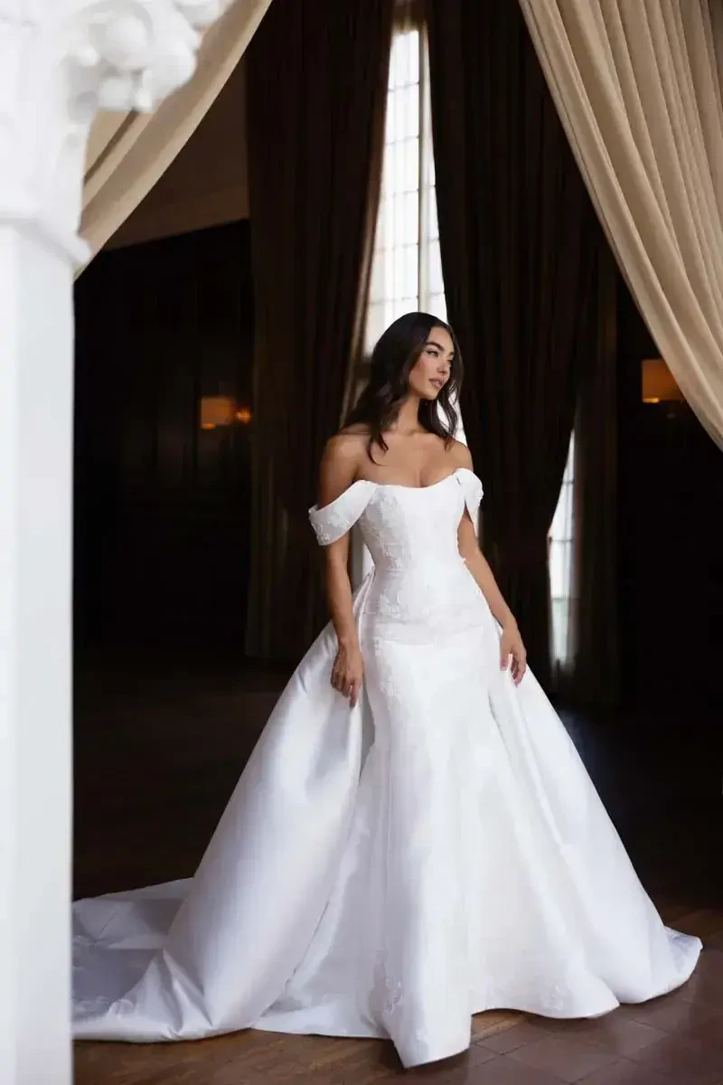 A woman in an elegant off-the-shoulder white wedding dress standing in a grand room with window light.