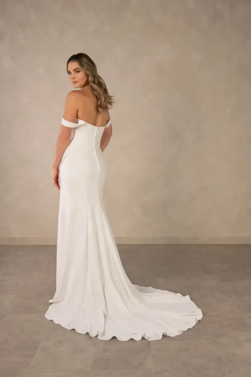 A woman in a white off-the-shoulder wedding dress, posing with her back to the camera, against a neutral background.