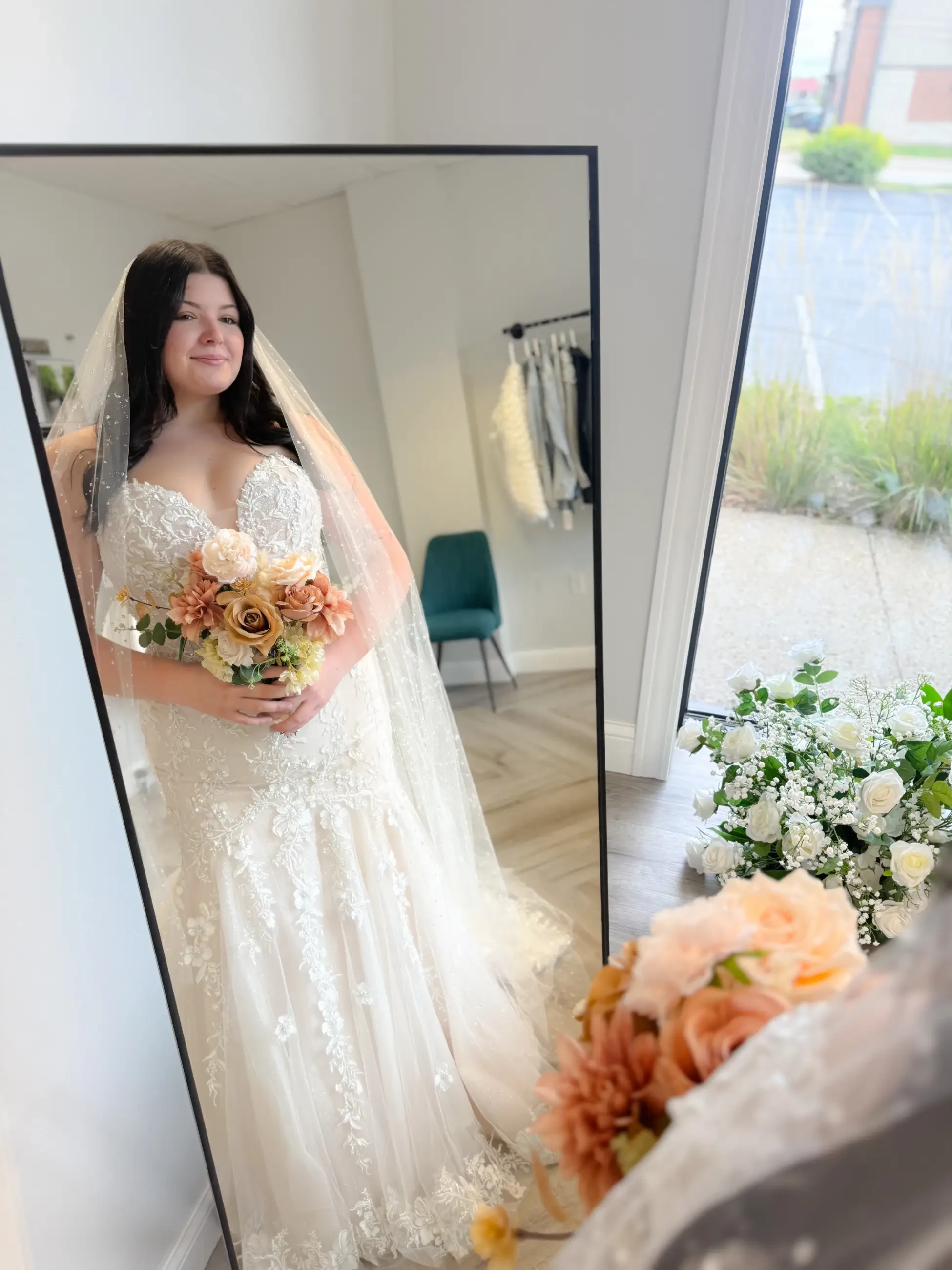 A bride in a beautiful wedding dress stands in front of a mirror, holding a bouquet of flowers. The reflection shows her smiling, and there is floral decor on the floor.
