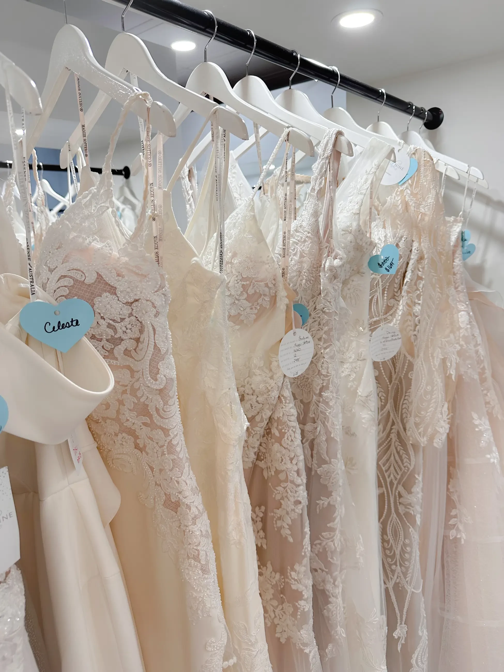 A row of elegant wedding dresses hanging on white hangers, each with a tag indicating selection options. The dresses feature various styles and designs, predominantly in soft, neutral colors.