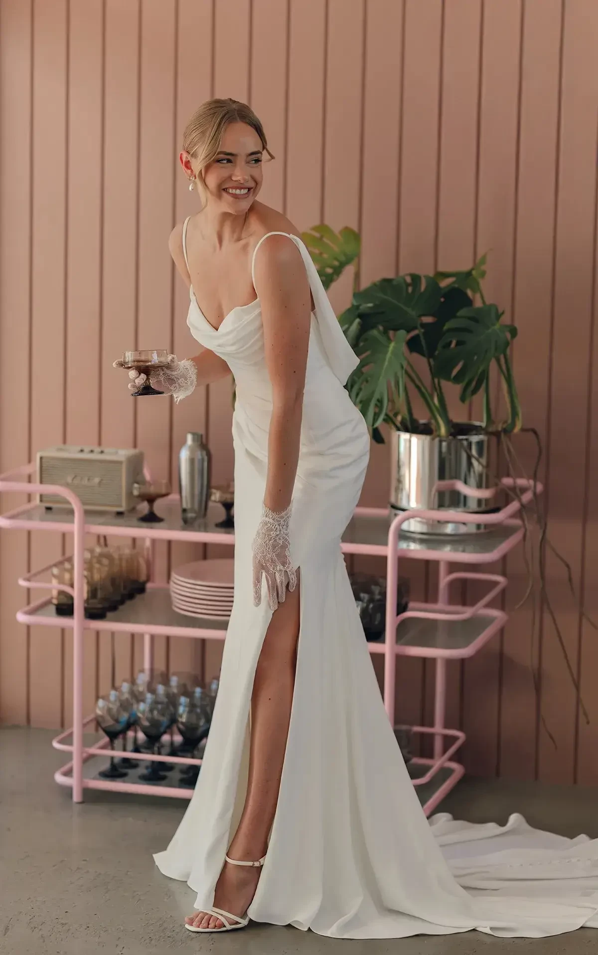 A woman in a white wedding dress with a slit stands smiling, holding a drink and wearing elegant gloves. Behind her is a pink wall and a bar cart with plants and decorative items.