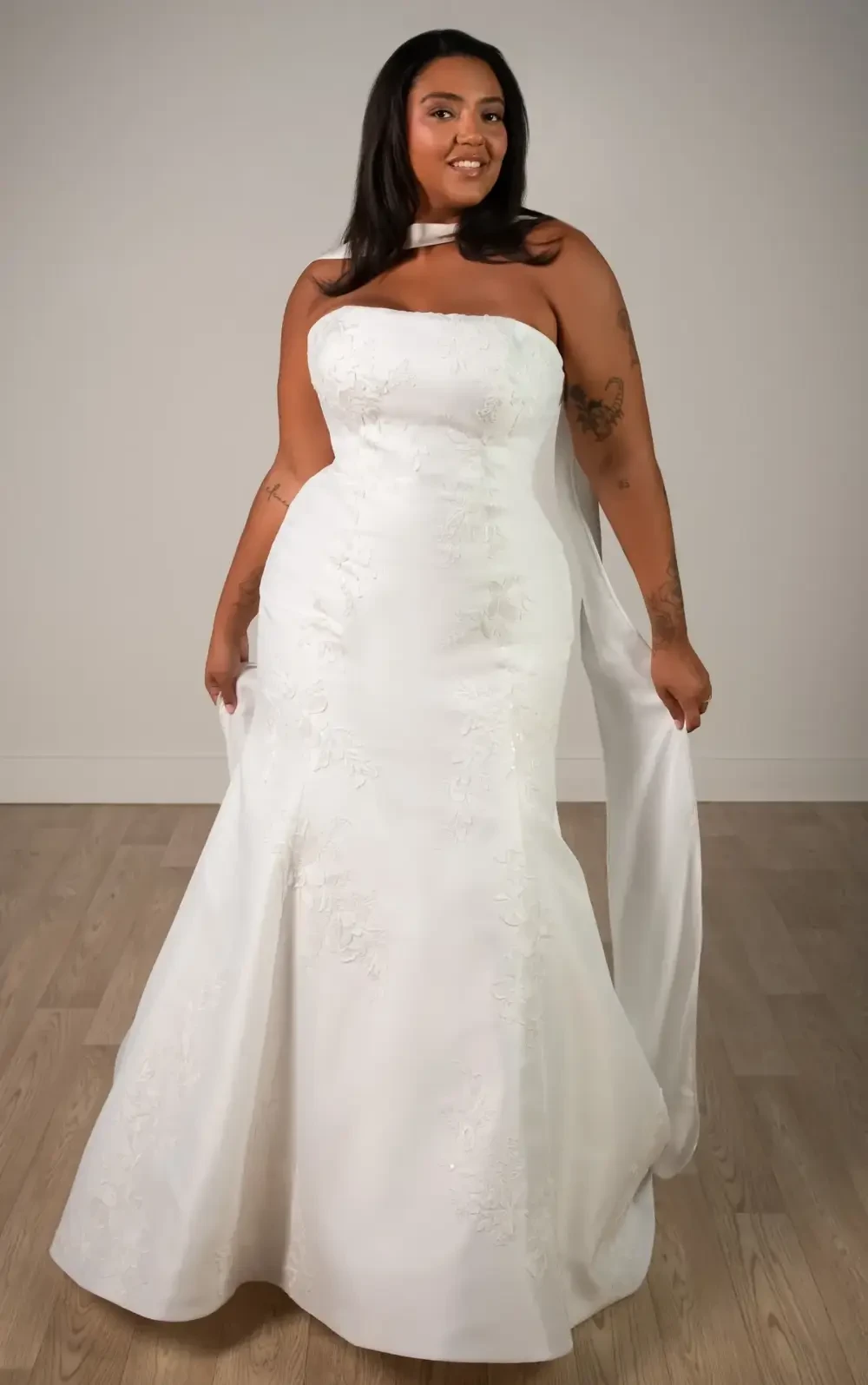 A person wearing a strapless white wedding dress with intricate detailing, standing confidently with a smile.